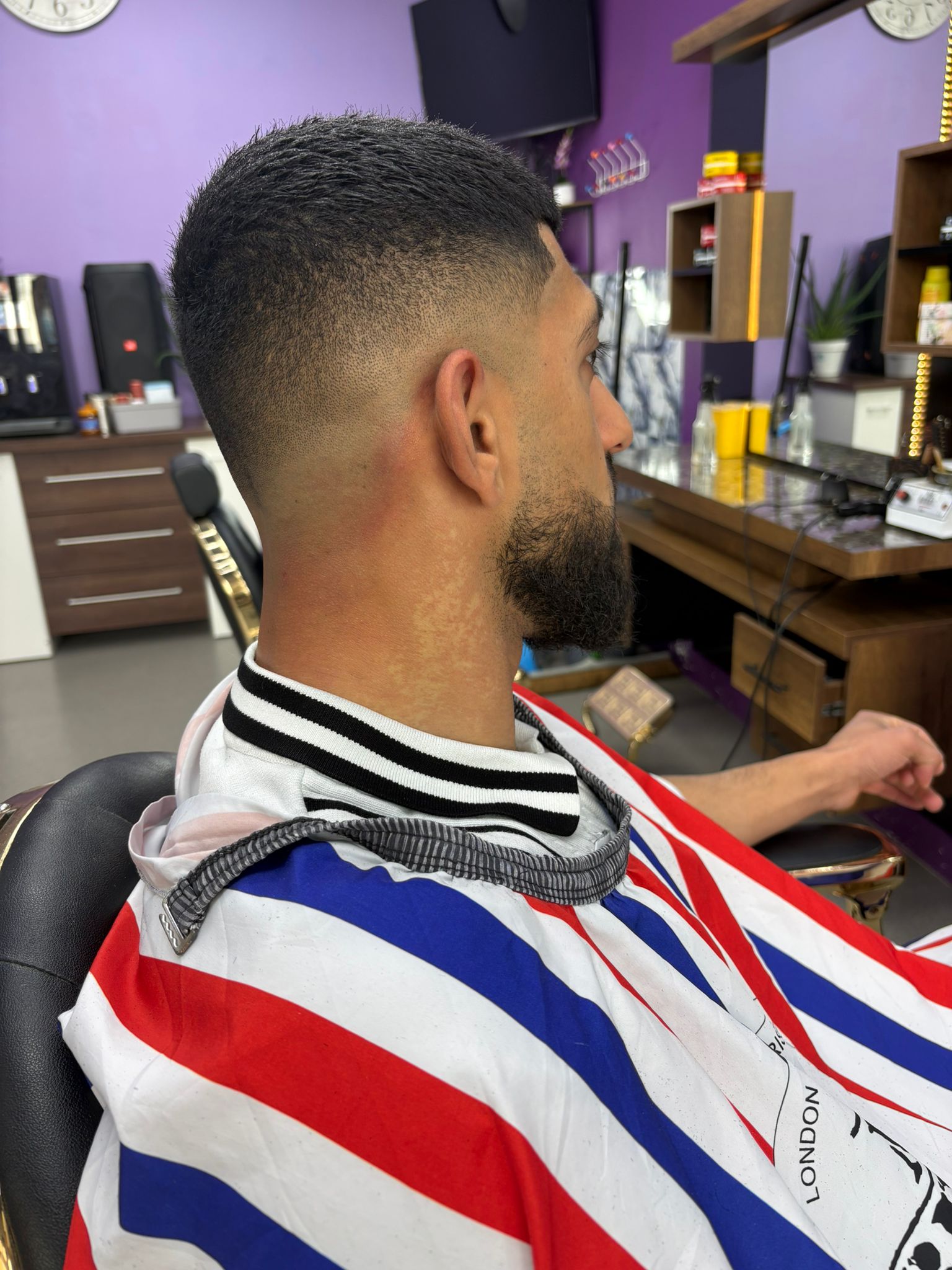 Klant in de stoel met een skin fade en een bijgewerkte baard, onder een gestreepte kapmantel